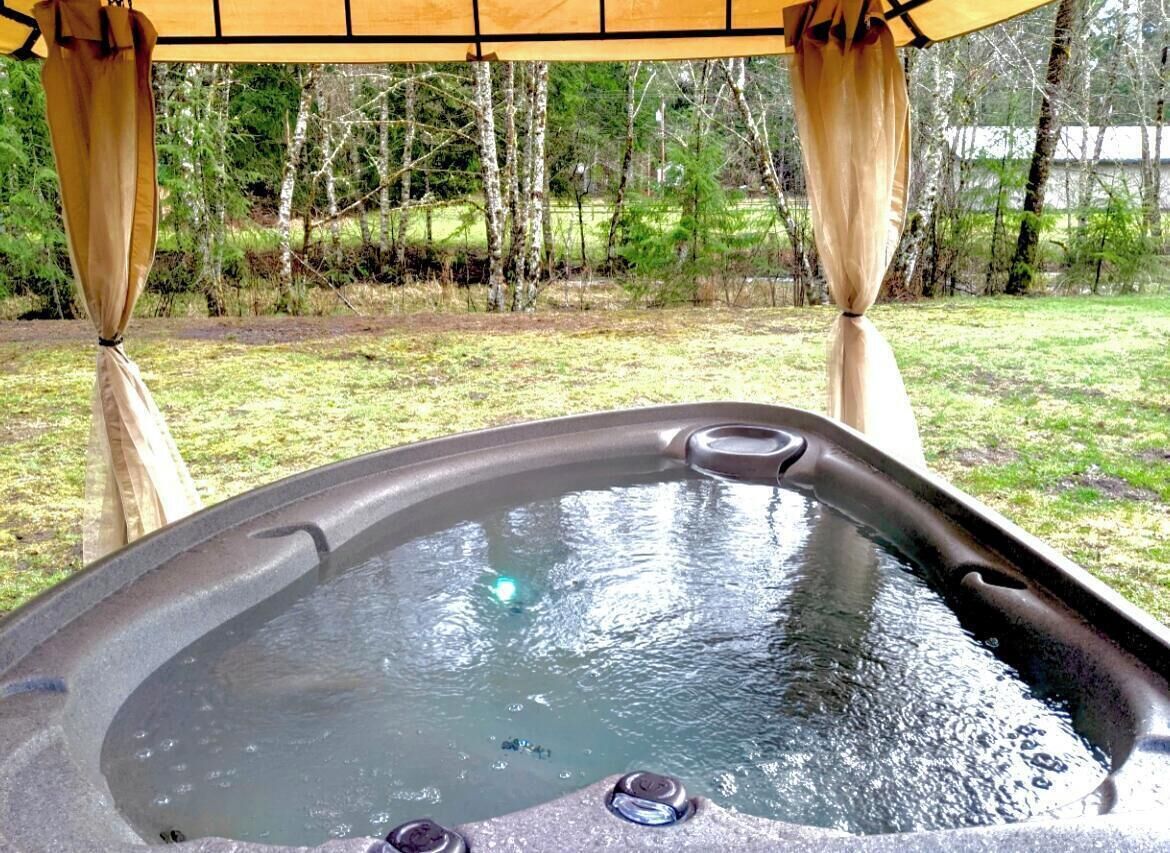 A hot tub under a wooden pergola with curtains, surrounded by a grassy yard and trees.