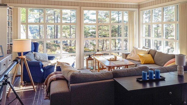 Ein gemütliches Wohnzimmer mit einem Ecksofa, einem Couchtisch und einem Esstisch. Große Fenster bieten einen Ausblick ins Freie.