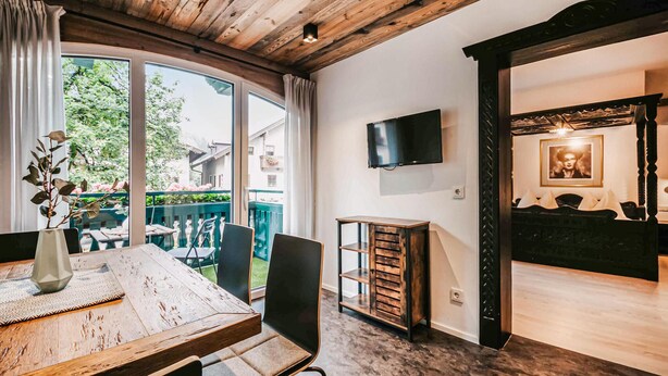 A dining area with a wooden table and chairs, a vase with a plant, and a view of the outside. - Ferienwohnung - Wagrain (Wagrain)