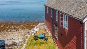 Ett rött trähus med ett tak av sten som vetter ut mot en stilla sjö eller ett annat stilla vattendrag.