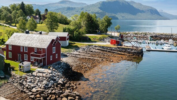 En röd byggnad med en grund av sten, en brygga med båtar och en bergig bakgrund.