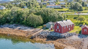 Ett hus vid kusten med en röd fasad, beläget nära en klippig strand och en vattenyta.
