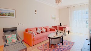 Una sala de estar con un sofá rosa, una mesa de centro de vidrio, un tapete estampado y un comedor con una mesa y sillas.