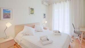 Una cama bien hecha con sábanas blancas, una mesita de noche con una lámpara, un escritorio con una silla y dos cuadros en la pared.