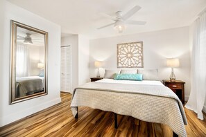 A bedroom with a large bed, wooden flooring, a ceiling fan, two bedside lamps, a decorative wall piece, and a mirror on the wall.