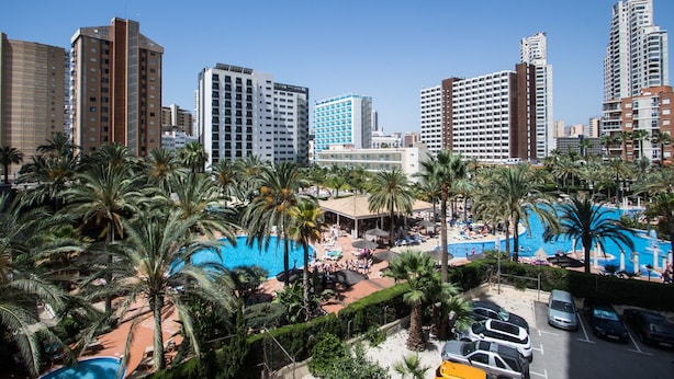 Un hôtel de grande hauteur doté d’une piscine sur le toit, entourée de palmiers, et d’un espace de stationnement.
