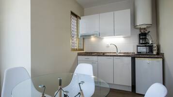 Una cocina compacta con armarios blancos, una mesa de comedor de vidrio y una cafetera.