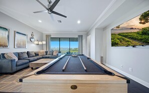 Une table de billard dans une pièce dotée d’un ventilateur de plafond, d’un canapé et d’une grande fenêtre offrant une belle vue.