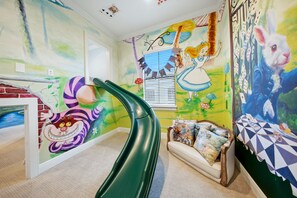 Une salle de jeux pour enfants avec un toboggan, des peintures murales colorées et un canapé agrémenté de coussins à motifs.