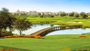 Un parcours de golf avec un pont enjambant un étang, entouré de pelouses parfaitement entretenues et bordé d’une rangée de maisons en arrière-plan.