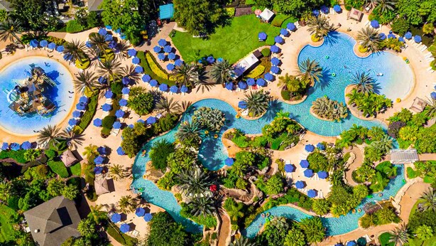Vue aérienne d’un complexe hôtelier doté de plusieurs piscines, de palmiers et de chaises longues.