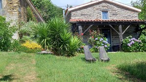 Uma casa de pedra com telhado de telhas, dois lounges na varanda frontal e diversas plantas e flores ao redor.