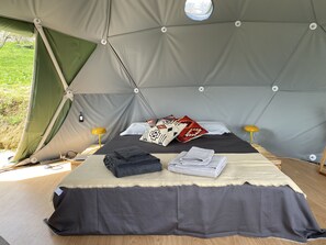 A tented room with a bed, pillows, folded towels, and a small table with a lamp.