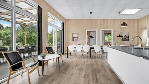 Een moderne keuken met houten lambrisering, een eethoek met witte stoelen en grote ramen met uitzicht op groen.