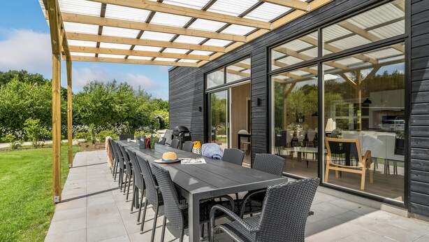 Een buitenpatio met een lange tafel en stoelen, een barbecue en een glazen deur naar een binnenruimte.