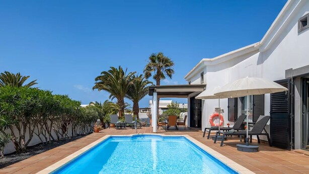A swimming pool with lounge chairs and umbrellas, surrounded by palm trees and a white building.