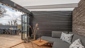 An outdoor seating area with a wooden deck, a glass door leading to a patio, and a grey sofa with floral cushions. - 3 bedroom beautiful home in Sydals (Sydals)