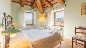 Ein Schlafzimmer mit einem Bett, zwei Vorhängen an den Fenstern, einer Nachttischlampe, einem Sessel und einer Holzbalkendecke.
