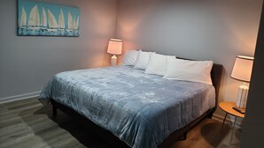 A bedroom with a bed, two bedside lamps, a painting of sailboats, and a small round table with a remote control. - Harrison Hotel Unit D
Pet Friendly 
Ground Level Unit (Garden City Beach)