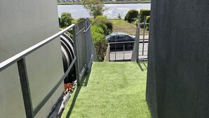 En balkon med kunstgræs, udsigt til vand og en parkeringsplads i baggrunden.