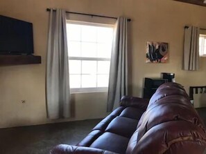 A living room with a dark brown sofa, a flat-screen TV mounted on the wall, and a window with curtains. - Big Bend Lodging at Ten Bits Ranch | Room #6 (Texas Hattery) (Terlingua)