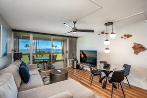 Una sala de estar moderna con un televisor grande, piso de madera, ventilador de techo y vistas al exterior.