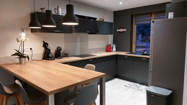 Une cuisine moderne avec une table en bois, des chaises noires, des luminaires suspendus et un réfrigérateur.