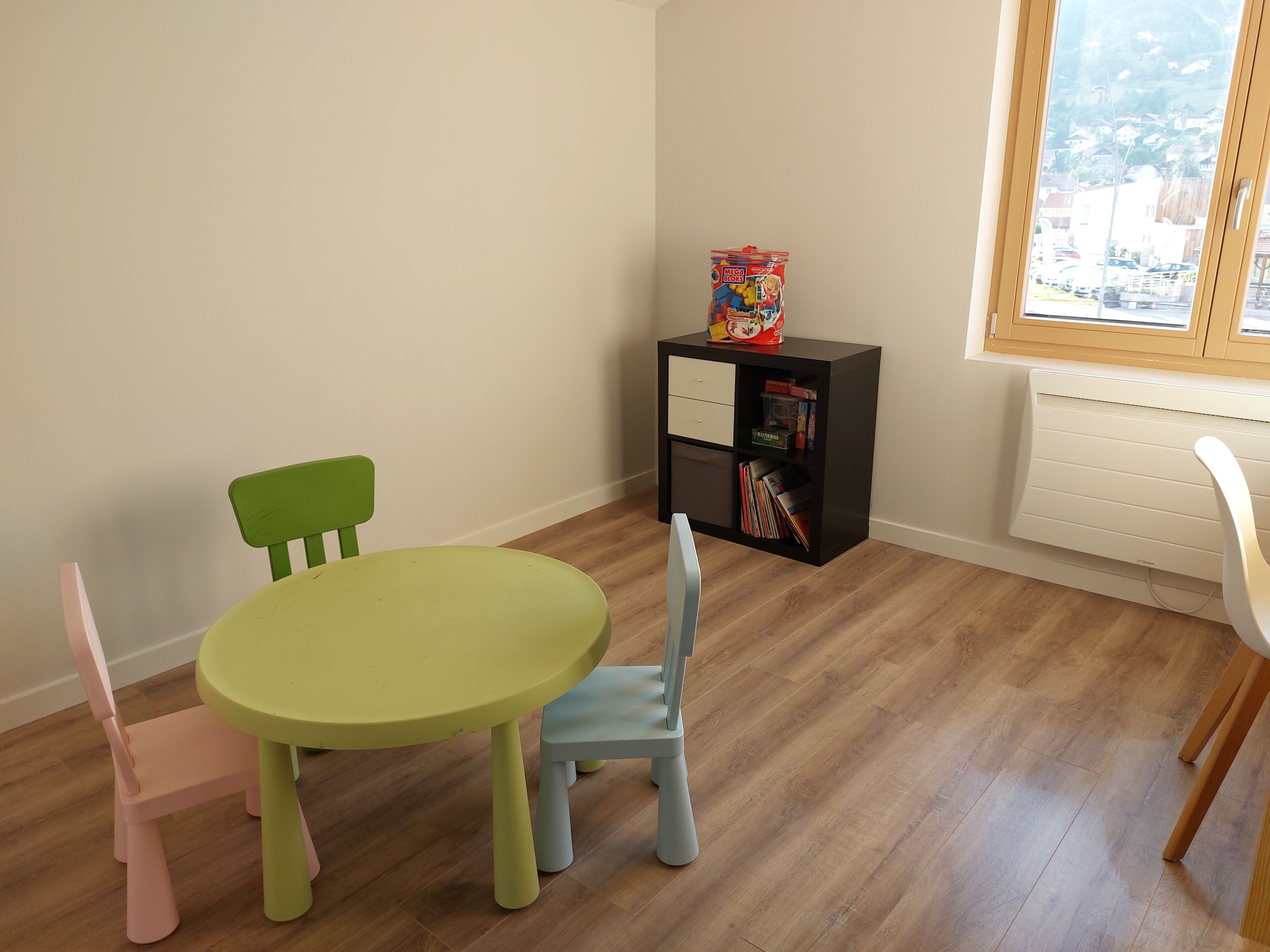 Una habitación con una mesa redonda, dos sillas, una estantería y una ventana con vistas a edificios.
