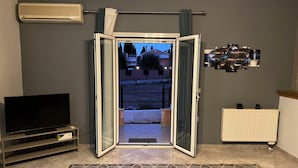 En åben dør fører ud til en balkon med udsigt over en bygning og træer. Et fjernsyn står på et stativ til venstre, og en radiator er placeret til højre.