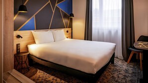 A modern hotel room with a large bed, a desk, and a chair. There is a window with curtains, a wall with geometric patterns, and a ceiling light. - Ibis Styles Valenciennes Petite Foret (Petite-Foret)
