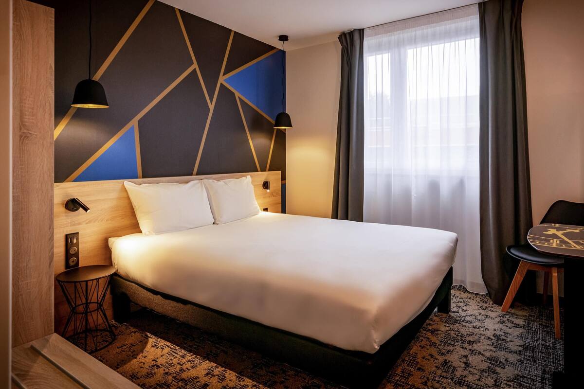 A modern hotel room with a large bed, a desk, and a chair. There is a window with curtains, a wall with geometric patterns, and a ceiling light.