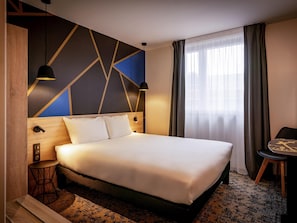 A modern hotel room with a large bed, a desk, and a chair. There is a window with curtains, a wall with geometric patterns, and a ceiling light. - Ibis Styles Valenciennes Petite Foret (Petite-Foret)