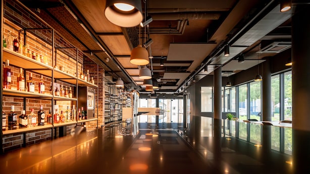 A spacious interior with a long counter displaying various bottles, large windows, and hanging pendant lights.