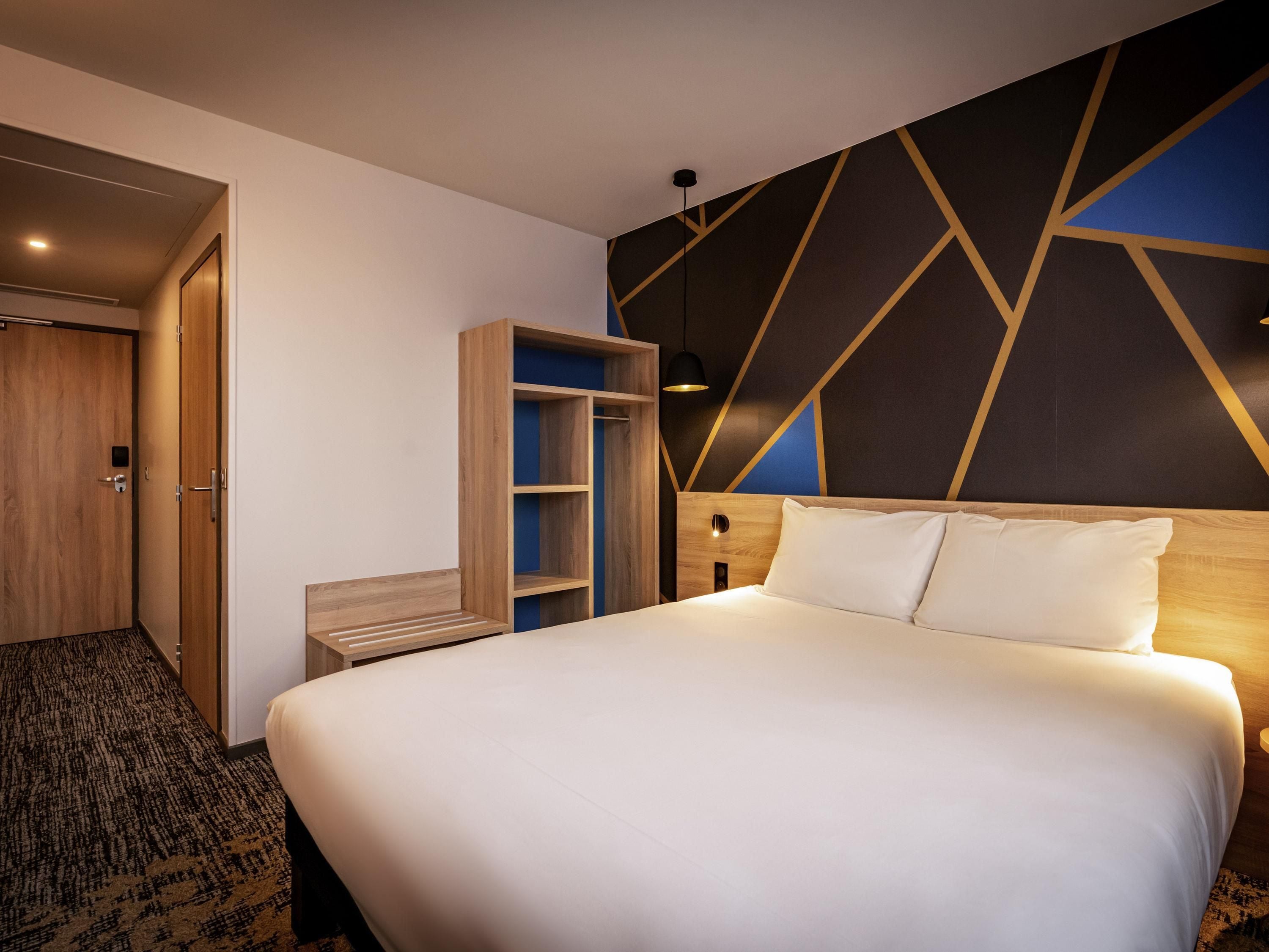 Hotelkamer met een groot bed, een kledingkast, een bankje en een wand met een geometrisch patroon.