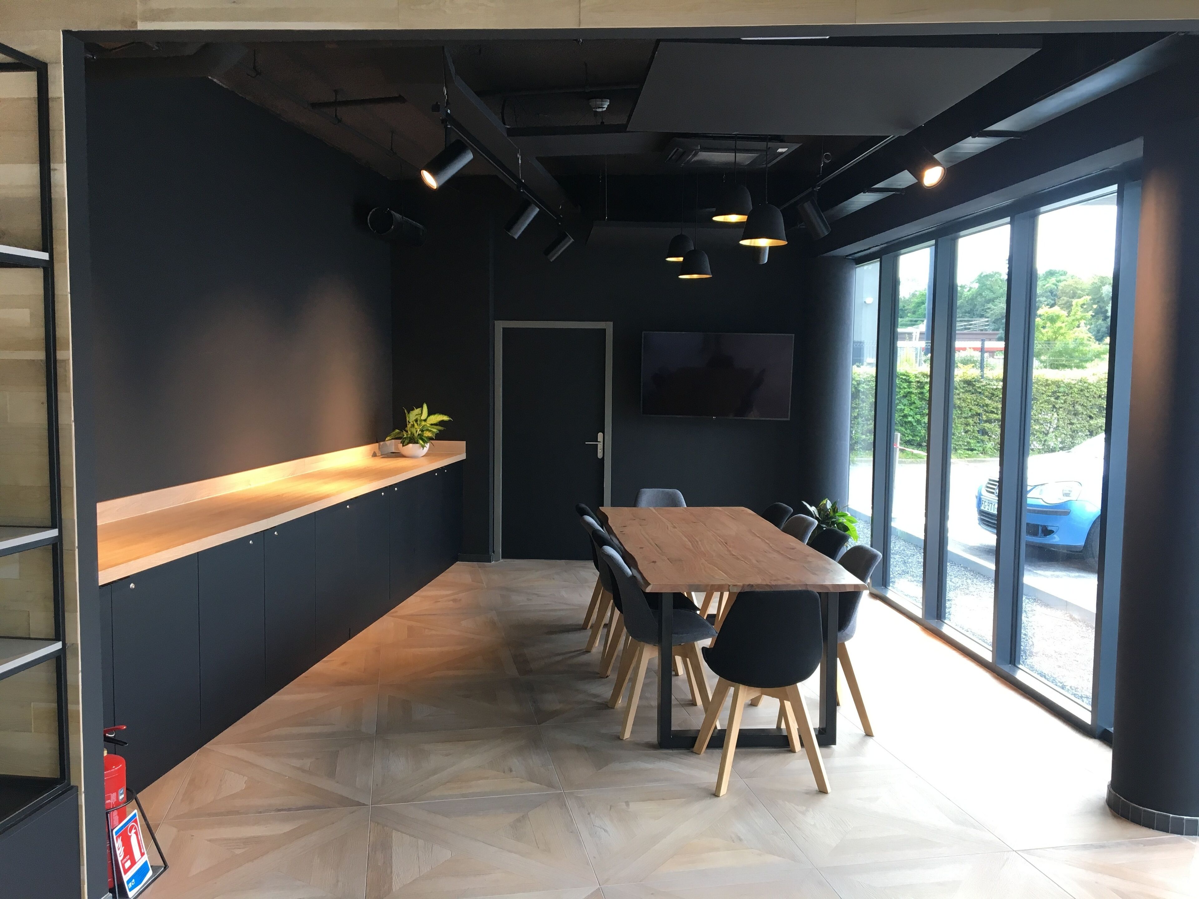 A modern conference room with a long wooden table, black chairs, a potted plant, and a large window offering a view of the outdoors.