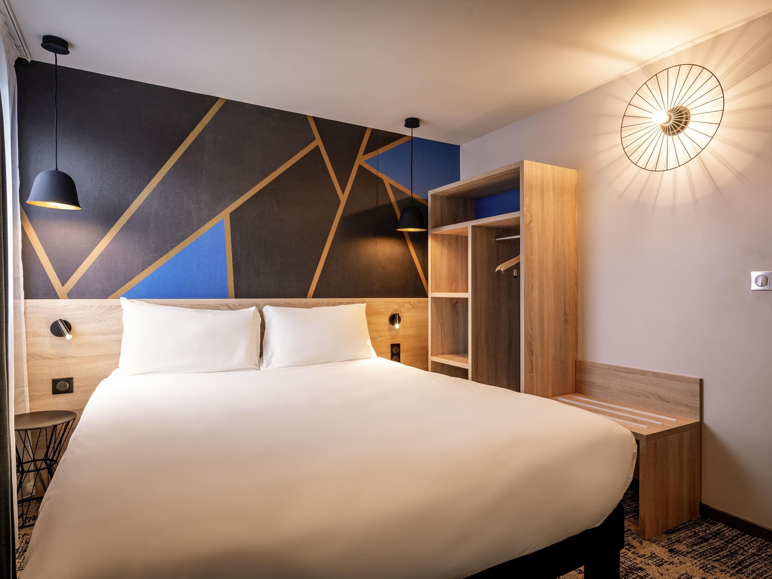 A modern hotel room with a large bed, built-in wardrobe, and a unique geometric wall design.