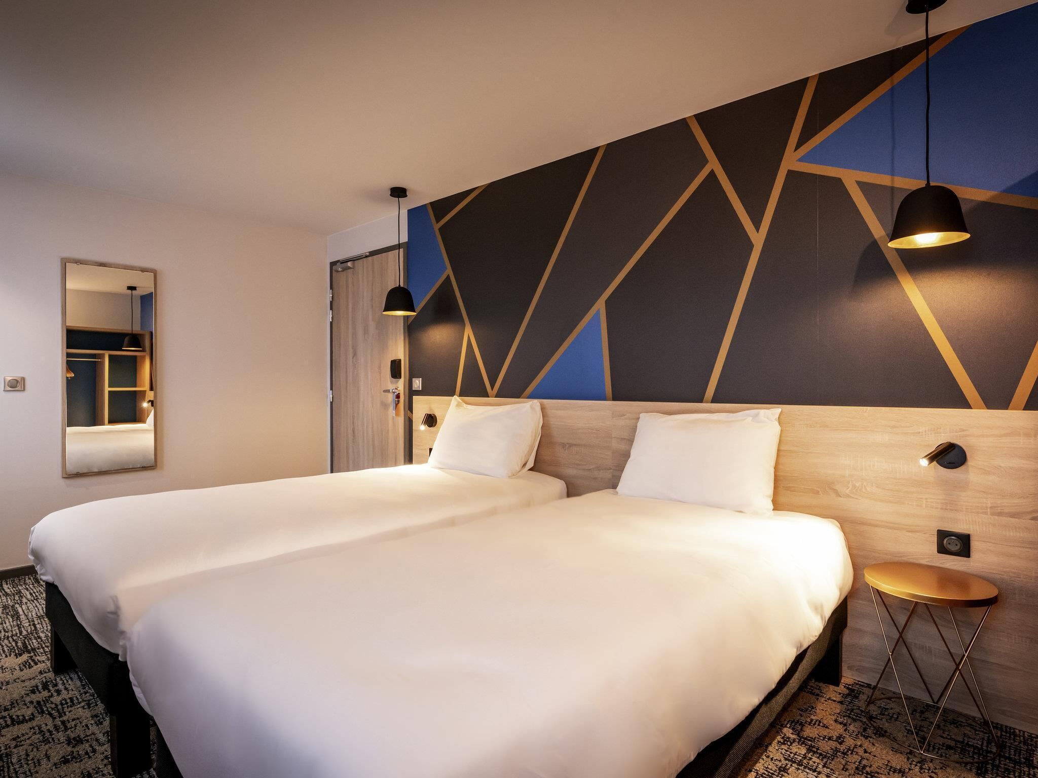 A double bed with a wooden headboard, a small round table, a mirror, and a geometric-patterned wall.