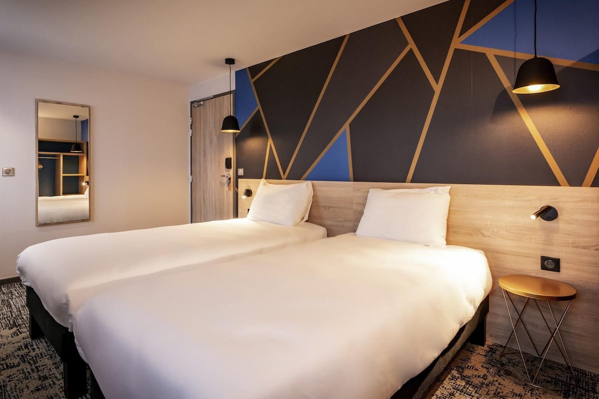 Un lit double avec une tête de lit en bois, une petite table ronde, un miroir et un mur orné d’un motif géométrique.