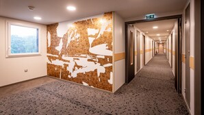 Pasillo de hotel con alfombra estampada, un gran muro abstracto y una ventana con vista a árboles.