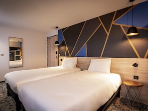 A double bed with white linens, a wooden headboard, a geometric-patterned wall, a small round side table, and a mirror on the wall. - Ibis Styles Valenciennes Petite Foret (Petite-Foret)