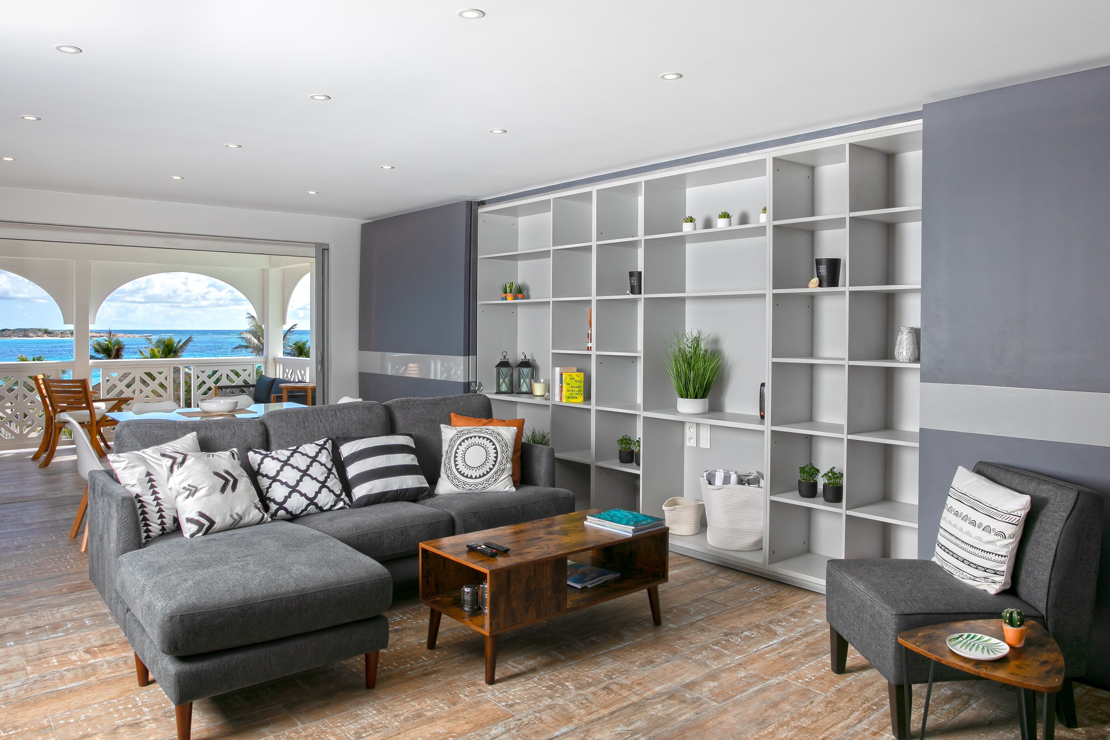 A modern living room with a grey sofa, a wooden coffee table, and a grey armchair. There is a white shelving unit against the wall with various decorative items.
