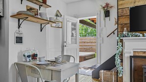 A dining area with a table set for two, a white chair with 'UTENSILS' labeled, and a wooden shelf above holding plates and a sign.