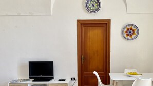 A room with a wooden door, a TV on a white cabinet, a white chair, and a small table with a bowl. - DAMMUSO to REKALE (Rekhale)