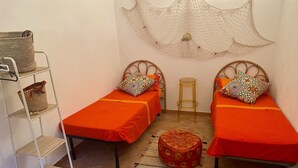 A small, cozy room with two single beds, a small stool, a shelf with baskets, and a decorative wall hanging. - DAMMUSO to REKALE (Rekhale)