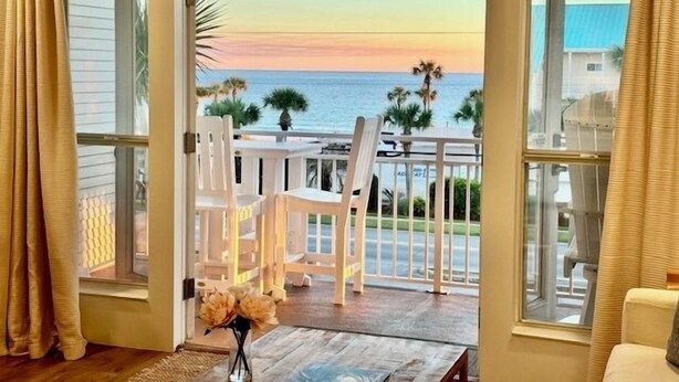 Une pièce offrant une vue sur l’océan au coucher du soleil, avec une table et des chaises en bois, un vase rempli de fleurs et un balcon doté d’une balustrade.