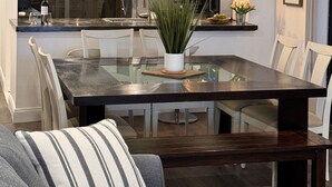 Un comedor moderno con una mesa de vidrio, sillas blancas y un banco. Hay lámparas colgantes sobre la mesa y una planta en maceta sobre ella.
