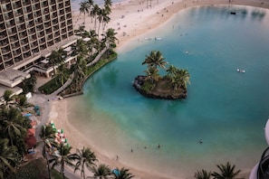A hotel with multiple floors, a beach, clear blue water, and palm trees.