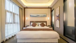 A hotel room with a large bed, a headboard with a painting, two wall-mounted lamps, and a large window with curtains.