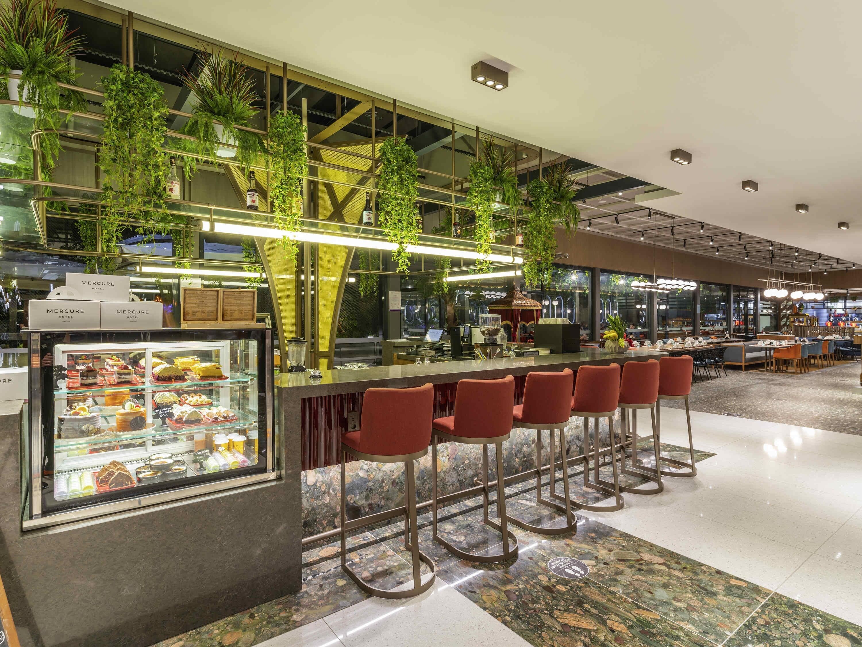 Un espace bar moderne avec un présentoir en verre rempli de desserts, des tabourets de bar rouges et des plantes suspendues.