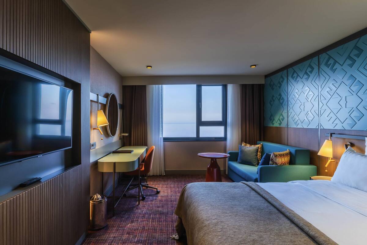 Ein Hotelzimmer mit Bett, Schreibtisch mit Stuhl, Fernseher, kleinem runden Tisch und Blick ins Freie.